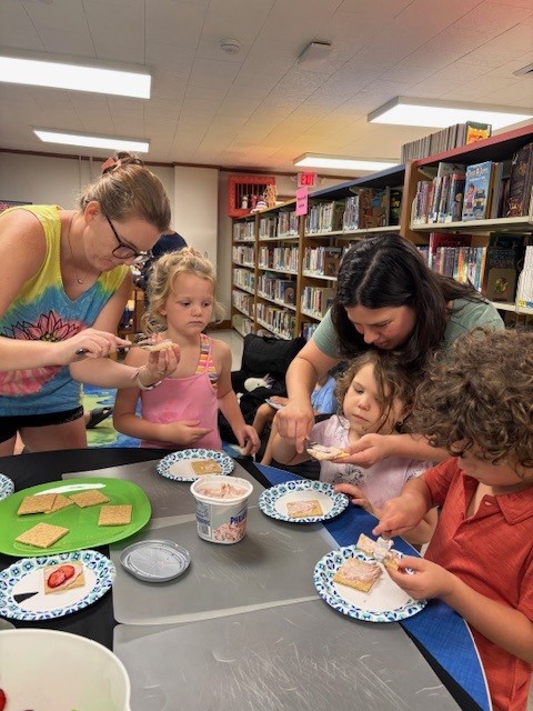 Children and adults making snacks with graham crackers and cream cheese in a library setting.